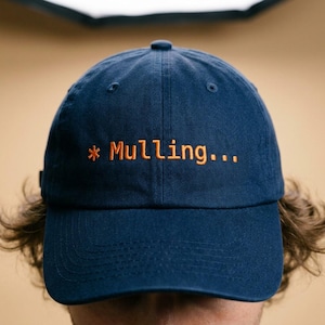May include: Navy blue baseball cap with the embroidered text "* Mulling..." in orange. The cap has a curved brim and a classic six-panel design. A casual accessory for everyday wear.