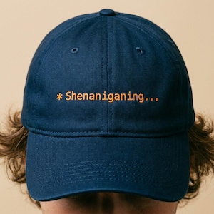 May include: A navy blue baseball cap with the text "* Shenanigans..." embroidered in orange. The cap has a curved brim and a button on top. The hat is a casual accessory.