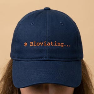 May include: A navy blue baseball cap with the text "* Bloviating..." embroidered in orange. The cap has a curved brim and a button on top. The hat is made of a soft material.