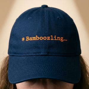May include: A navy blue baseball cap with the word "*Bamboozling..." embroidered in orange thread. The cap has a curved brim and a button on top. The hat is made of a soft, durable fabric.