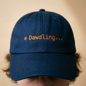 May include: Navy blue baseball cap with the embroidered text "* Dawdling..." in orange. The cap has a curved brim and a button on top. A casual accessory.