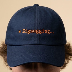May include: Navy blue baseball cap with the embroidered text "* Zigzagging..." in orange. The cap has a curved brim and a button on top. The hat is made of a soft material.