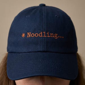 May include: Navy blue baseball cap with the word "Noodling..." embroidered in orange thread. The cap has a curved brim and a rounded top. A casual accessory for everyday wear.