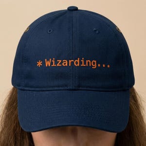 May include: A navy blue baseball cap with the text "* Wizarding..." embroidered in orange. The cap has a curved brim, a button on top, and adjustable strap. The cap is made of a durable fabric.