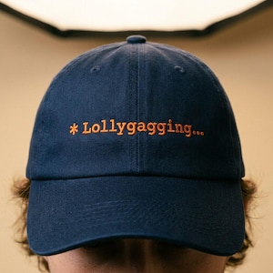 May include: A navy blue baseball cap with the word "Lollygagging..." embroidered in orange thread across the front. The cap has a curved brim and a classic six-panel design. The background is a soft, neutral color.