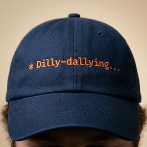 May include: Navy blue baseball cap with the embroidered text "* Dilly-dallying..." in orange. The cap has a curved brim and a button on top. The hat is made of a soft fabric.