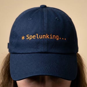 May include: Navy blue baseball cap with the word "* Spelunking..." embroidered in orange thread. The cap has a curved brim and a button on top. A casual accessory for outdoor enthusiasts.
