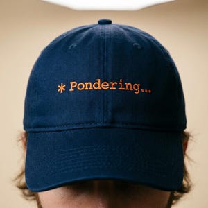May include: A navy blue baseball cap with the embroidered text "* Pondering..." in orange. The cap has a curved brim and a rounded top. The hat is made of a soft fabric.