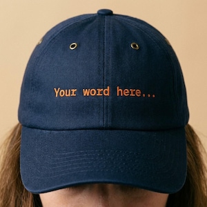 May include: Navy blue baseball cap with the embroidered text "Your word here..." in orange. The cap has a curved brim, a top button, and two small eyelets on each side. A casual accessory.
