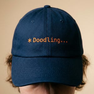 May include: A navy blue baseball cap with the word "* Doodling..." embroidered in orange thread. The cap has a curved brim and a classic six-panel design, suitable for casual wear.