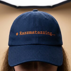 May include: A navy blue baseball cap with the embroidered text "* Razzmatazzing..." in orange. The cap has a curved brim and a classic six-panel design. The hat is made of a soft fabric.