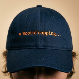 May include: Navy blue baseball cap with the embroidered text "*Bootstrapping..." in orange. The cap has a curved brim and a button on top. A casual accessory.