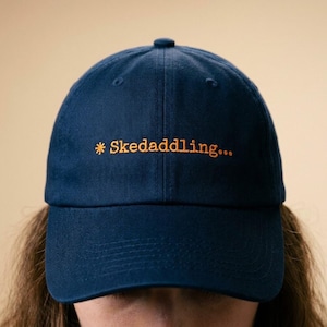 May include: Navy blue baseball cap with the embroidered text "* Skedaddling..." in orange. The cap has a curved brim and a rounded crown. The hat is made of a soft, durable fabric.