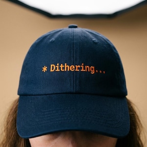 May include: A navy blue baseball cap with the embroidered text "* Dithering..." in orange. The cap has a curved brim and a structured crown. The background is a solid tan color.