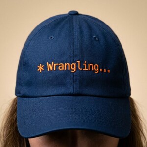 May include: A navy blue baseball cap with the embroidered text "* Wrangling..." in orange. The cap has a curved brim and a button on top. The hat is made of a soft, durable fabric.