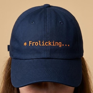 May include: Navy blue baseball cap with the embroidered text "* Frolicking..." in orange. The cap has a curved brim and an adjustable strap at the back. The hat is made of a soft, durable fabric.