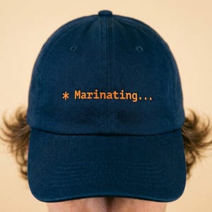May include: A navy blue baseball cap with the text "* Marinating..." embroidered in orange. The cap has a curved brim and a classic six-panel design. The hat is made of a soft, durable fabric.