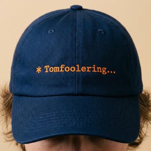 May include: Navy blue baseball cap with the embroidered text "* Tomfoolering..." in orange. The cap has a curved brim and a button on top. The hat is a classic style, suitable for casual wear.