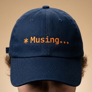 May include: Navy blue baseball cap with the word "* Musing..." embroidered in orange. The cap has a curved brim and an adjustable strap at the back. The hat is made of a soft, durable fabric.