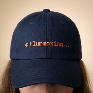 May include: Navy blue baseball cap with the word "*Flummoxing..." embroidered in orange thread across the front. The cap has a curved brim and a button on top. The background is a solid, neutral color.