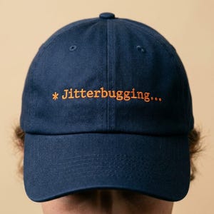 May include: Navy blue baseball cap with the text "* Jitterbugging..." embroidered in orange. The cap has a curved brim and a button on top. The hat is made of a soft fabric.