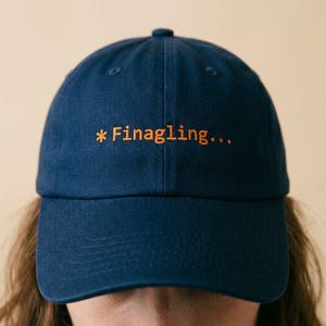 May include: A navy blue baseball cap with the text "*Finagling..." embroidered in orange thread. The cap has a curved brim and a button on top. The hat is a casual accessory.