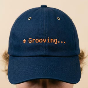 May include: A navy blue baseball cap with the text "* Grooving..." embroidered in orange. The cap has a curved brim and two small ventilation holes on the side.