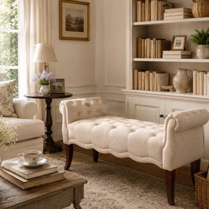 May include: A cozy living room scene featuring a cream-colored tufted bench with curved arms and dark wooden legs. A matching armchair, round side table, and a bookcase filled with books complete the elegant, neutral-toned decor. A coffee table holds books and a teacup.