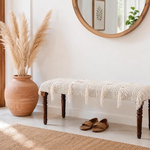 May include: A neutral-toned entryway scene featuring a bench with a fringed top and dark wooden legs. A large terracotta vase holds tall, dried pampas grass. Brown leather sandals sit near the bench. A round mirror and framed artwork are visible.
