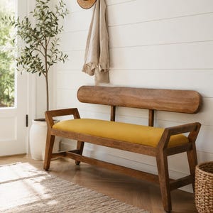 May include: Wooden bench with a yellow cushion. The bench has a curved back and armrests. A straw hat and a beige jacket hang on the white wall behind the bench. A small tree in a white pot is to the left.