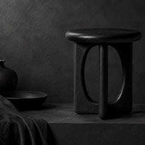 May include: A black wooden stool with a circular top and a base featuring four curved, open-arched legs. The stool is positioned next to a black ceramic vase and a shallow black bowl, all rendered in a monochromatic dark colour palette.