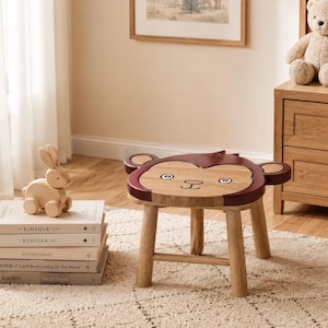 May include: A wooden stool featuring a monkey face design on the seat, with a natural wood finish and dark brown painted accents on the ears and head. The stool has four sturdy legs and a flat top surface with a simple, hand-drawn facial expression.