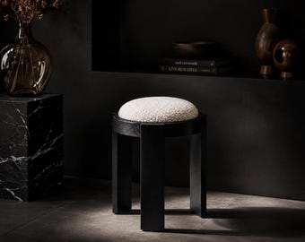 Puede incluir: Un taburete redondo con estructura de madera negra, cuatro patas rectas y un asiento circular tapizado en tela bouclé de color blanco roto con textura. El taburete presenta un diseño limpio y estructural con un asiento claro.