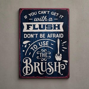 May include: A rectangular metal sign with a dark blue wood-grain background and white text that reads: IF YOU CAN'T GET IT - with a - FLUSH DON'T BE AFRAID TO USE - THE BRUSH. The sign features a graphic illustration of a toilet brush on the right side.