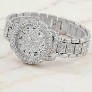 May include: A silver wristwatch with a sparkling, gem-studded face and strap. The watch features Roman numeral hour markers and a round face. The strap is made of rectangular links. The watch is set against a light background.