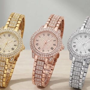 Op de afbeelding: Drie met strass versierde polshorloges in goud, roségoud en zilver. Elk horloge heeft een ronde wijzerplaat met Romeinse cijfers en een bijpassende met strass bedekte band. De horloges worden op een licht oppervlak weergegeven.