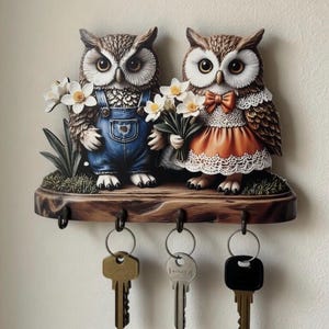 May include: A decorative key holder featuring two owl figurines holding flowers. The owls are perched on a wooden base with three key hooks. One owl wears blue overalls, and the other wears an orange dress. The keys hanging below are various shapes and colors.