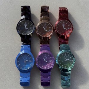 May include: Six metallic watches in various colors: black, bronze, red, blue, purple, and teal. Each watch has a round face with a date display and a matching metal band. The watch faces have a subtle design.