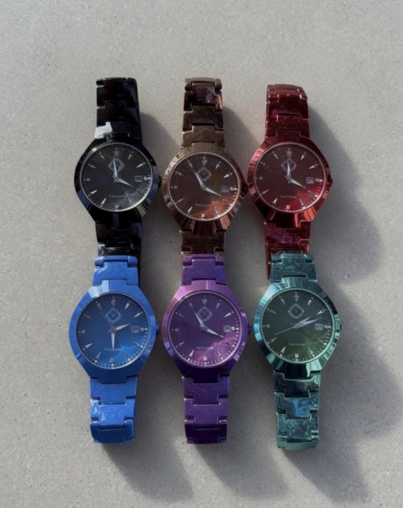 May include: Six metallic watches in various colors: black, bronze, red, blue, purple, and teal. Each watch has a round face with a date display and a matching metal band. The watch faces have a subtle design.