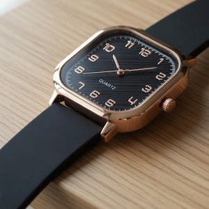 May include: A square-faced wristwatch with a black dial and rose gold-colored case. The watch has a black band and displays the word "QUARTZ" on the face. The watch is resting on a wooden surface.
