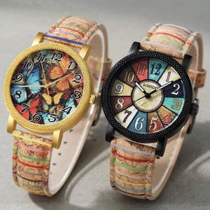 Könnte beinhalten: Zwei Armbanduhren mit einzigartigen Designs. Eine hat ein goldfarbenes Gehäuse und ein Zifferblatt voller bunter Schmetterlinge. Die andere hat ein schwarzes Gehäuse mit einem farbenfrohen nummerierten Zifferblatt. Beide Uhren haben Armbänder im Kork-Stil mit bunten Streifen.