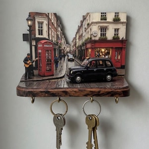 Puede incluir: Un llavero decorativo con una escena callejera londinense en miniatura con una cabina telefónica roja, un taxi negro y un músico. La repisa de madera tiene dos ganchos para llaves.