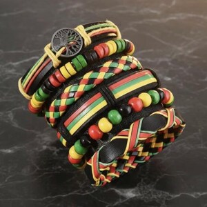May include: A stack of multi-layered bracelets featuring black leather bands, braided cords, and wooden beads in red, yellow, and green. One bracelet includes a circular metal charm depicting a tree silhouette with intricate branch details.