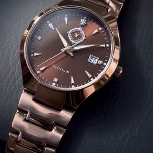 May include: A rose gold wristwatch with a brown face and matching band. The watch face features silver-colored hands and markers, a date display, and the brand name "Valirium". The watch is set against a dark background.