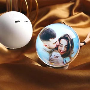 May include: Two spherical devices, one plain matte white with a visible charging port and speaker grille, and one featuring a high-resolution color photograph of a couple embracing. The photo-printed sphere has a smooth, glossy finish and a small attached cord.