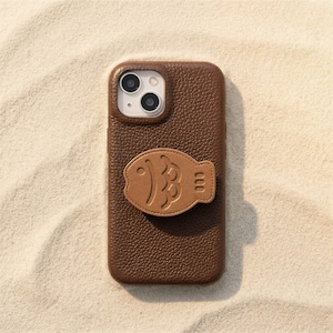 May include: A brown leather phone case with a fish-shaped grip. The case has a textured surface and a camera cutout. The fish grip is a lighter shade of brown and features embossed details. The phone case is resting on a sandy surface.