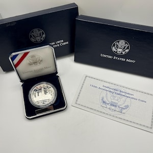 May include: A silver Proof Silver Dollar coin from the Smithsonian Institution's 150th Anniversary Commemorative Coins, displayed in a blue velvet case. The case is open, revealing the coin and a certificate of authenticity. Two navy blue boxes are in the background.