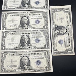 May include: Several vintage one-dollar silver certificates. Each bill features a portrait of George Washington and the text "The United States of America." The bills are in various conditions, with visible serial numbers and seals.