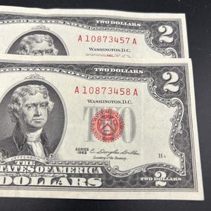 May include: Two United States two-dollar bills from the 1963 series. Each note features a portrait of Thomas Jefferson, the text "The United States of America", "Two Dollars", "Washington D.C.", and a red seal. Serial numbers A10873457A and A10873458A are visible.