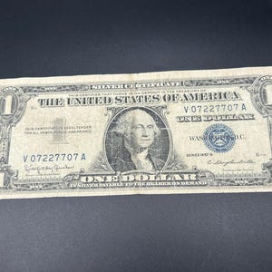 May include: A vintage one-dollar silver certificate from the United States of America. The bill features a portrait of George Washington, the text "ONE DOLLAR", and the serial number V07227707A. The bill is in shades of green and black.